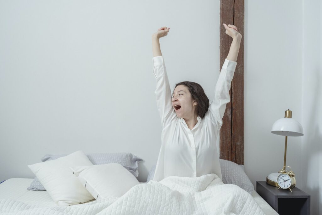Sleepy brunette sitting on white comfortable bed under blanket and stretching arms up while yawning and looking away during morning time at home