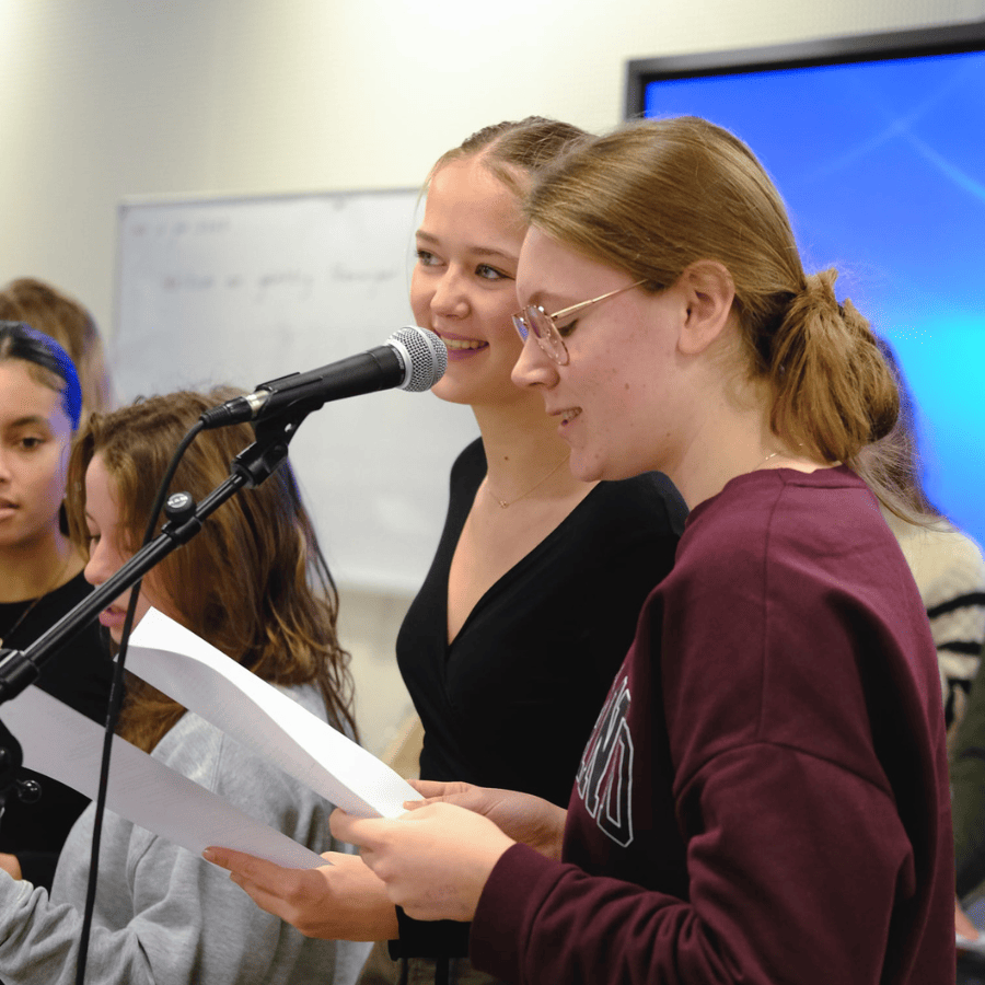 Twee bovenbouw leerlingen zingen lachend met blaadjes in hun handen achter een microfoon