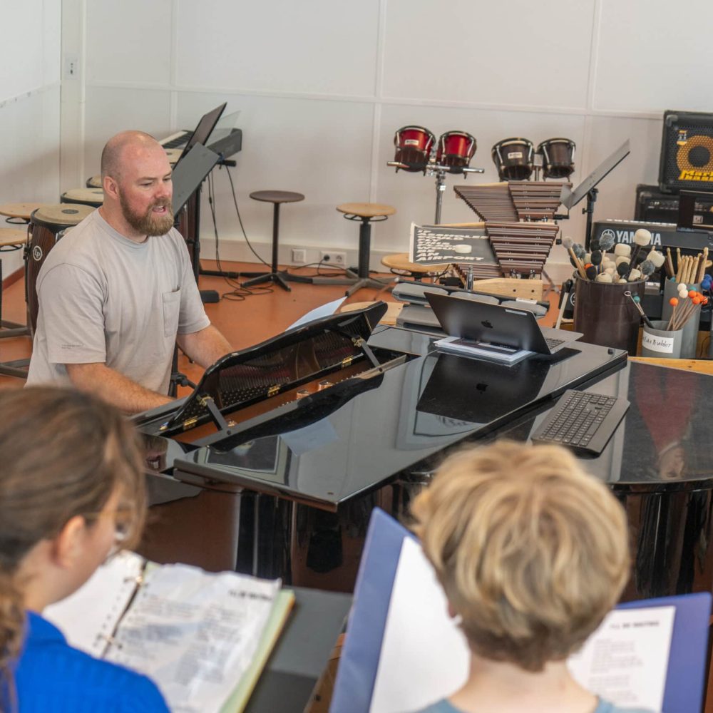 Muziekdocent Rudolf Draijer speelt vleugel in muzieklokaal tijdens muziekles, leerlingen luisteren aandachtig.